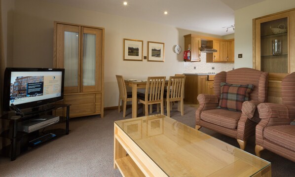A living room with a TV, wooden furniture, a dining table, and a kitchen area with wooden cabinets. - Macdonald Spey Valley Resort (Aviemore)