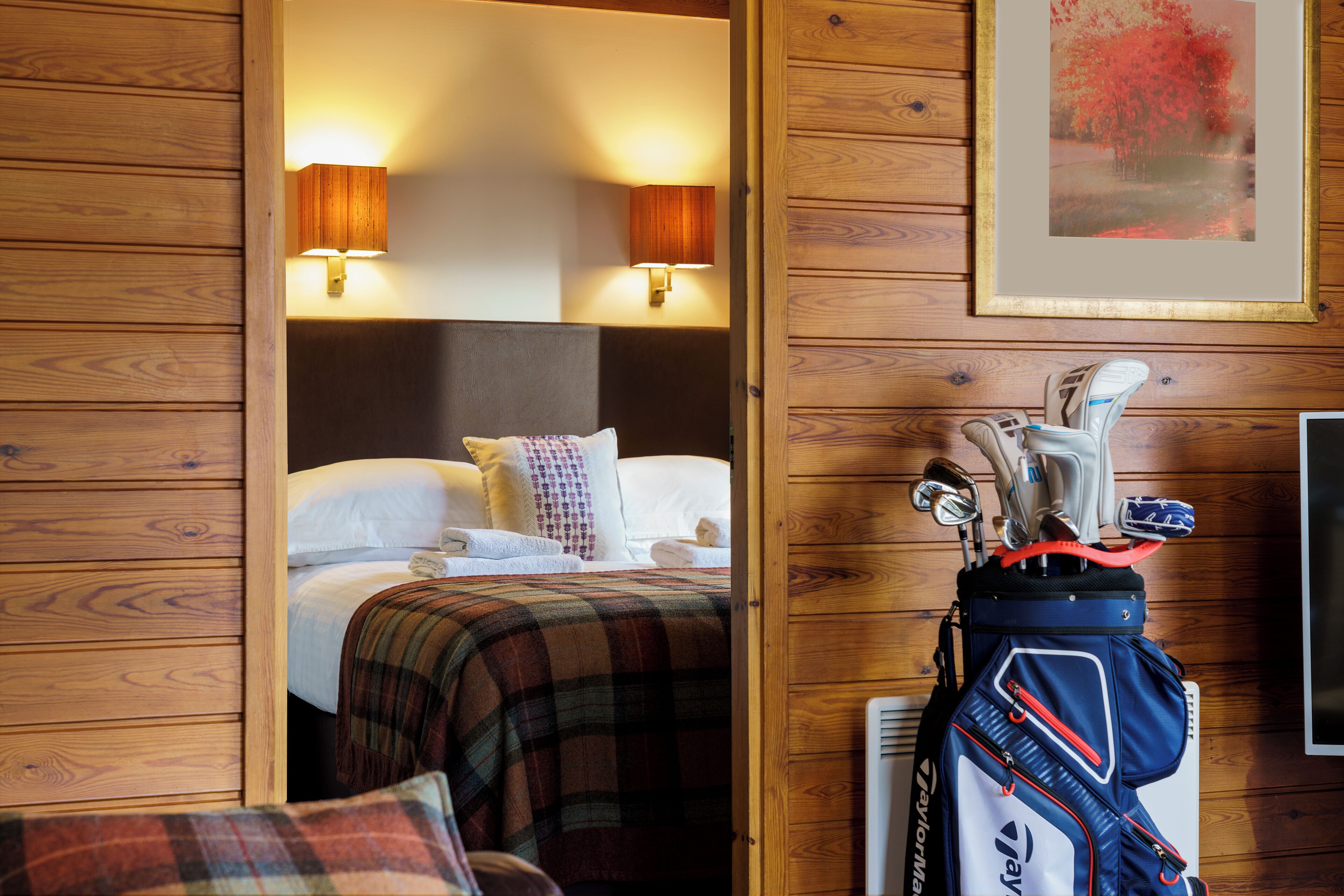 Une pièce aux murs lambrés, comprenant un lit, deux lampes fixées au mur, un tableau encadré et un sac de golf.