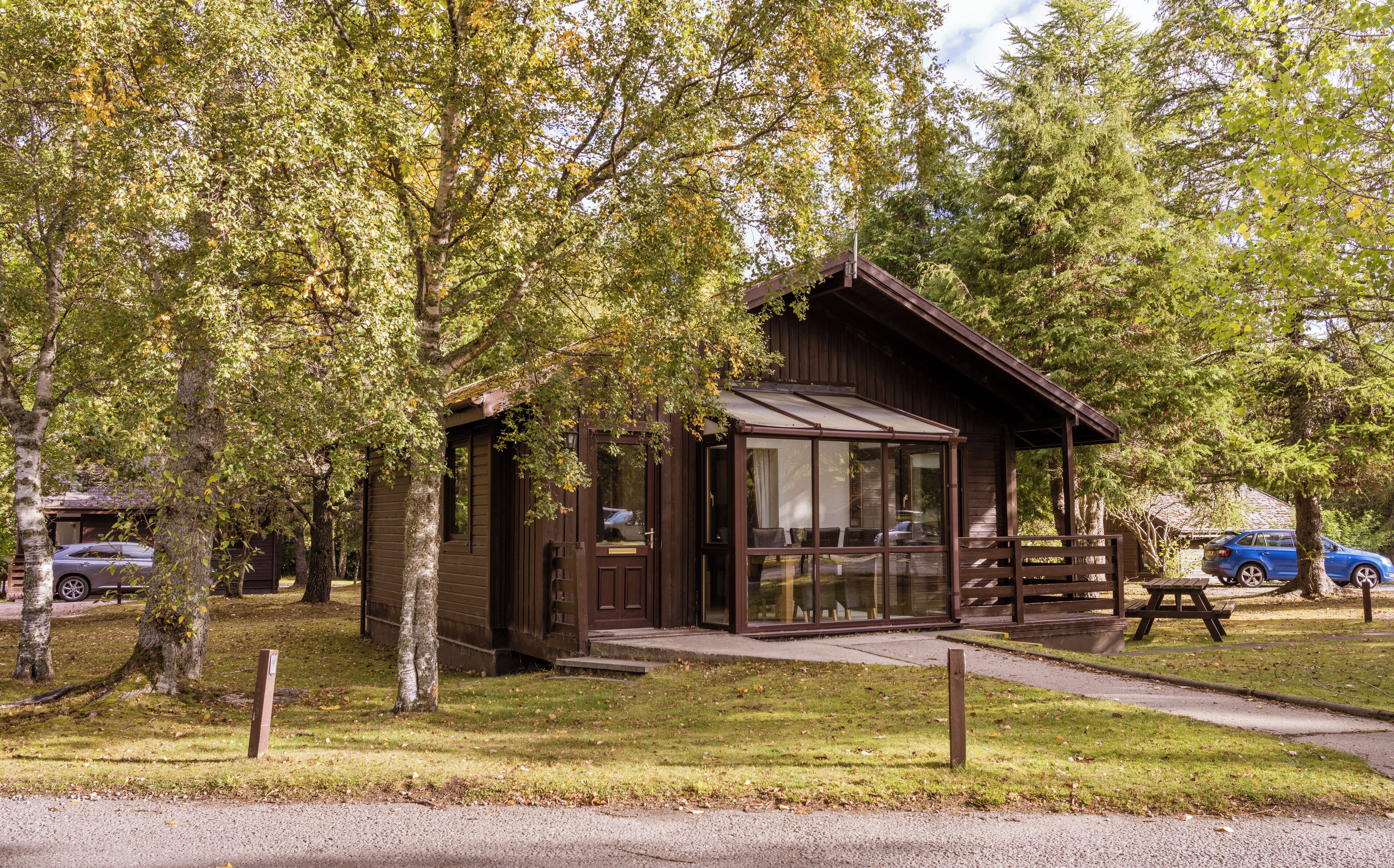 A small wooden cabin with a glass door, surrounded by trees and a picnic table.