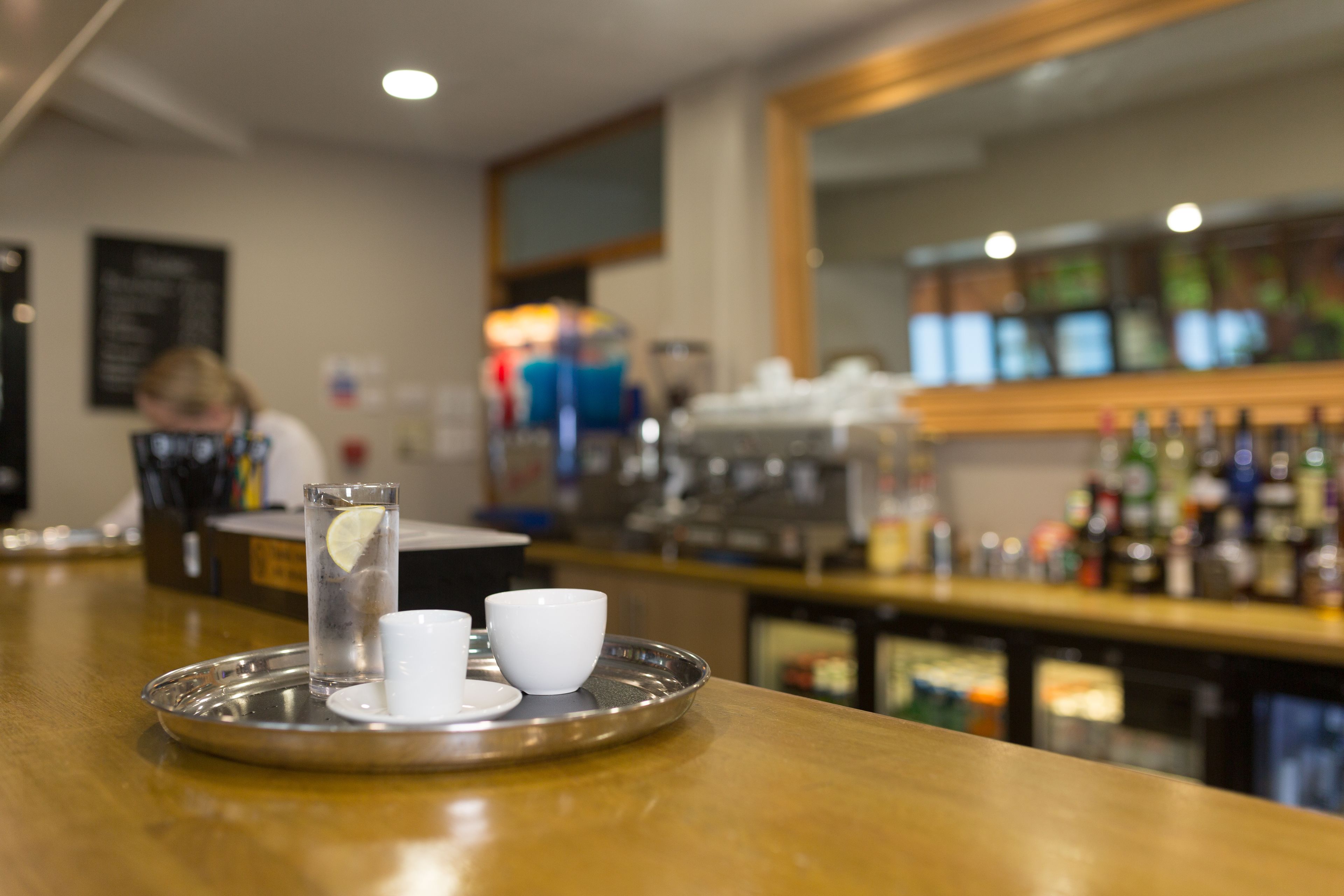 A bar area with a tray containing a glass of water, a white cup, and a saucer.