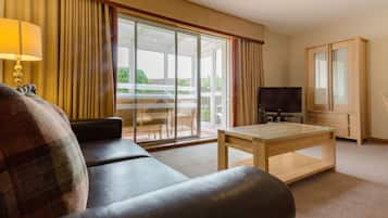 A hotel room with a sofa, a glass-top table, a television, and a sliding glass door leading to a balcony.