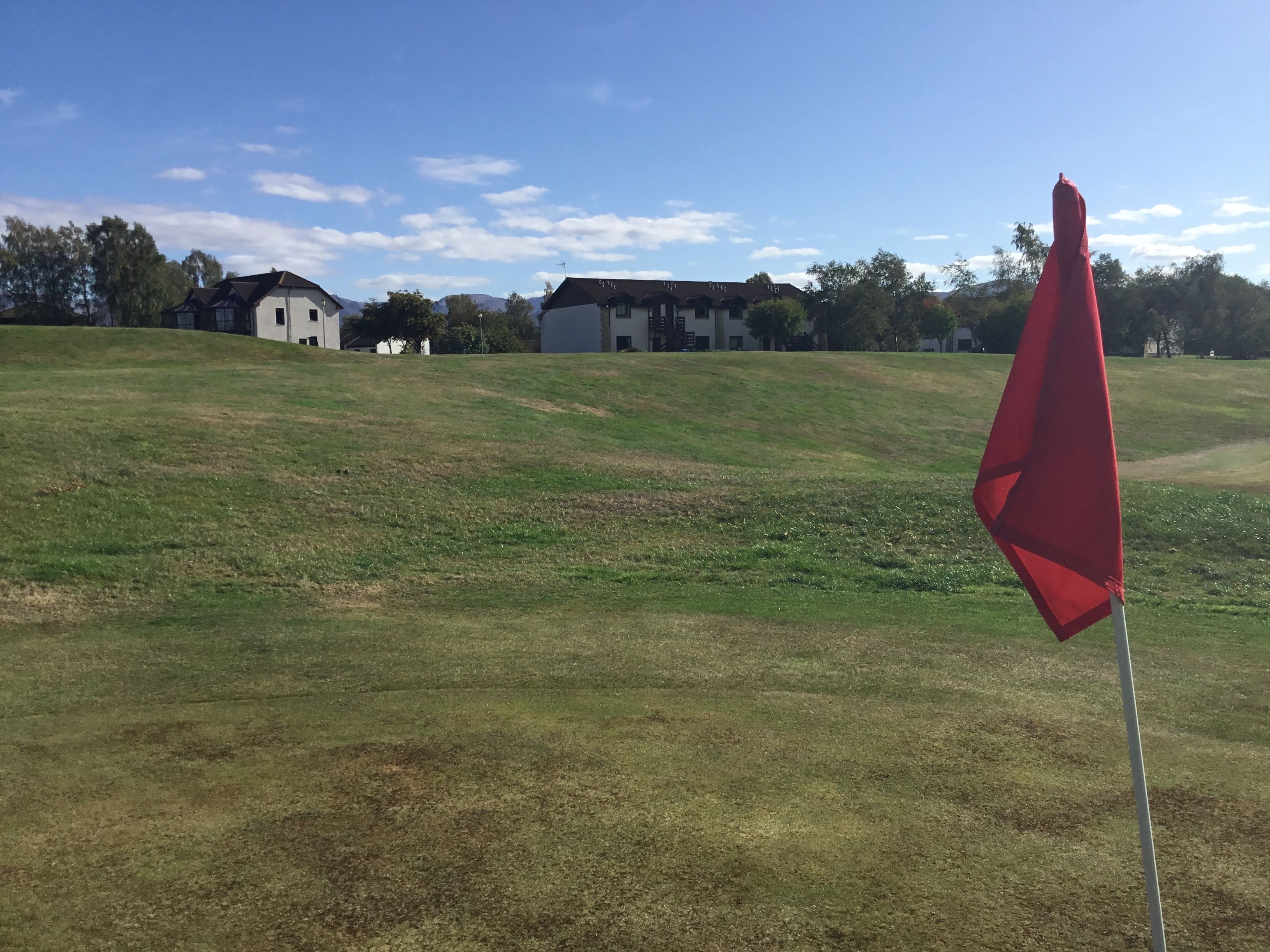 A golf course with a red flag on a pole, a grassy hill, and buildings in the background.
