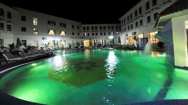 Vista nocturna de un patio de hotel con piscina, sillas de descanso y un edificio con varias ventanas.