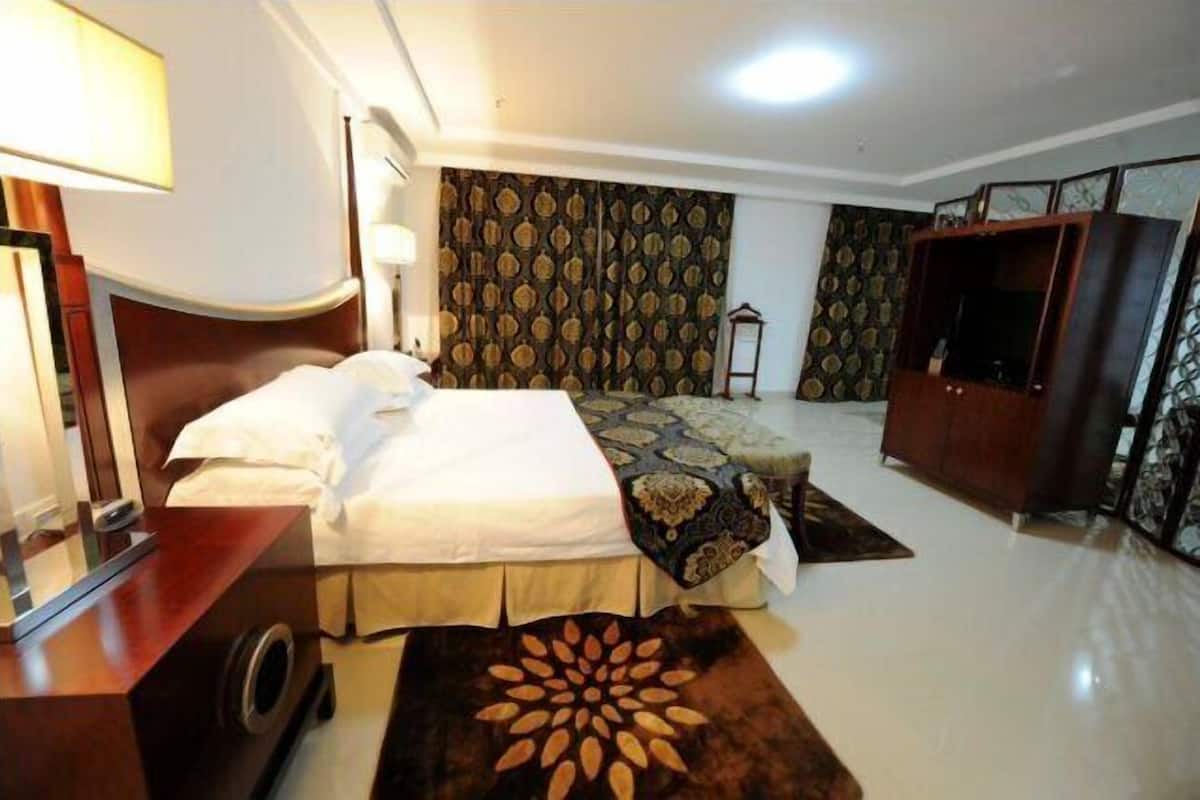 A hotel room with a large bed, a wooden wardrobe, a desk with a lamp, and patterned curtains.