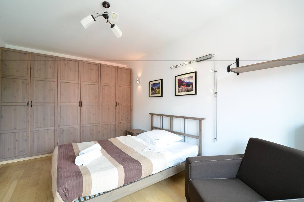 A bedroom with a bed, a sofa, wooden wardrobes, and two framed pictures on the wall.