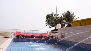 A swimming pool with water jets, surrounded by lounge chairs and a palm tree. - BWC Hotel (Lagos)