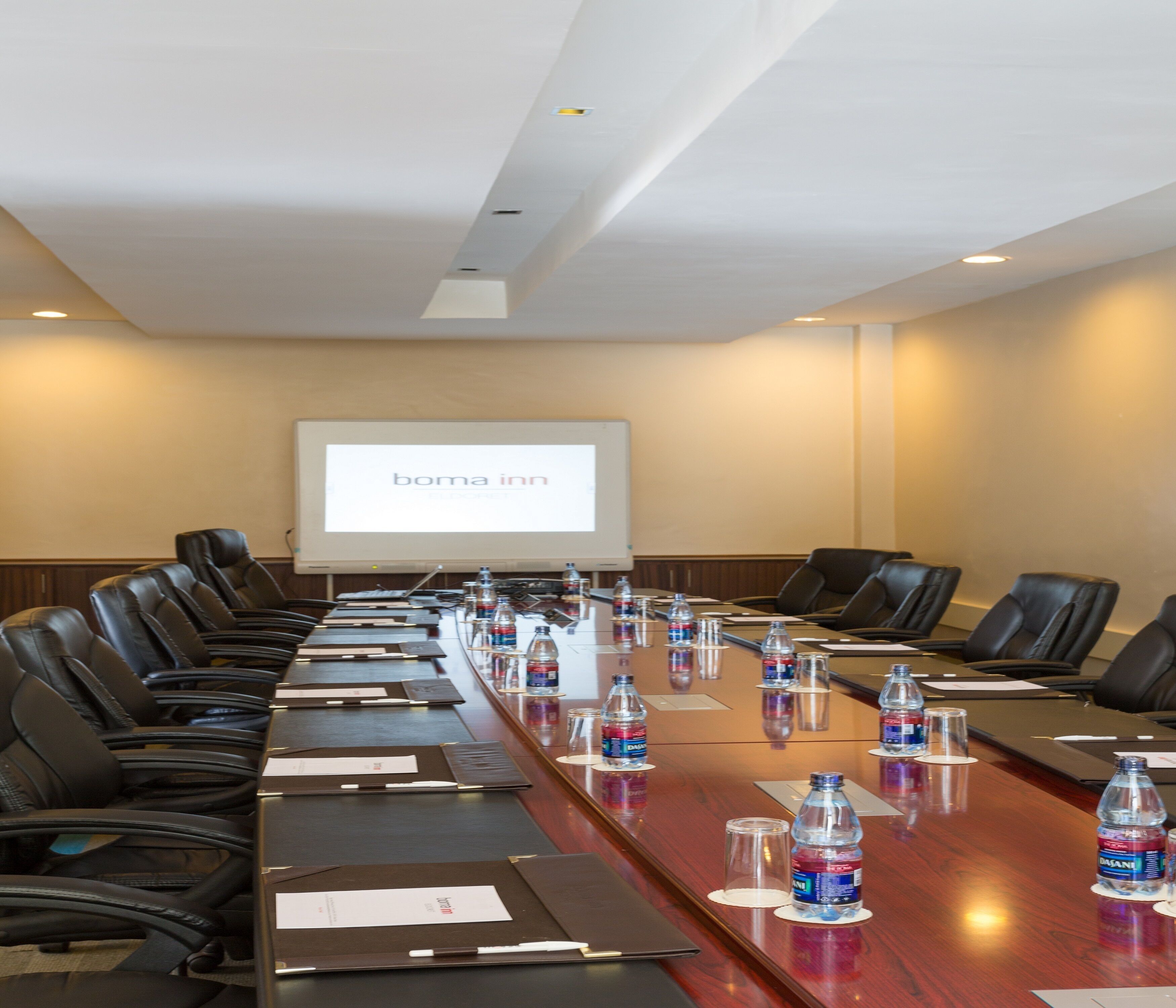 A conference room with a long table, black leather chairs, water bottles, and a projector screen displaying "boma inn".