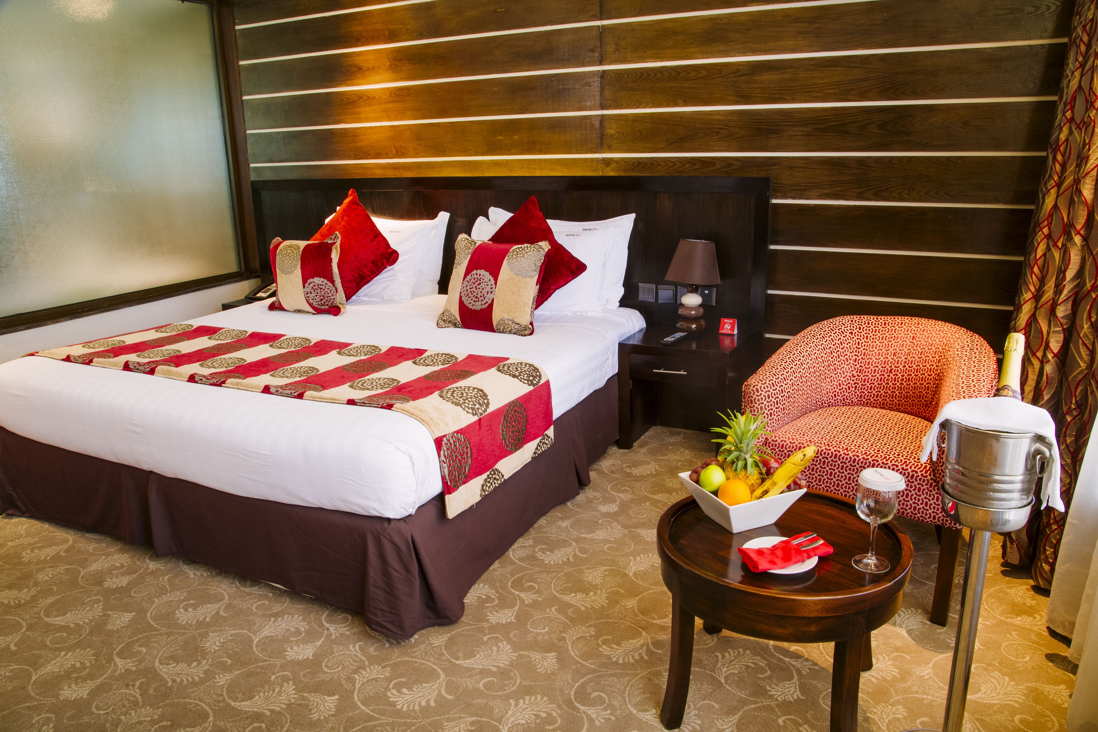A hotel room with a large bed, a nightstand, a red patterned armchair, a small table with fruit and a glass, and a minibar.