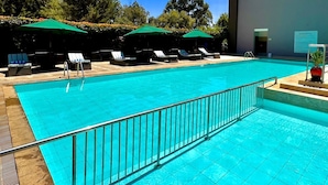 A swimming pool with lounge chairs and umbrellas, surrounded by trees and a building.