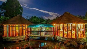 A thatched-roof structure with glass walls, surrounded by water, and set against a night sky.