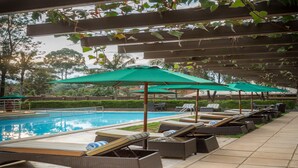 A poolside area with lounge chairs and green umbrellas, surrounded by greenery and a clear sky.