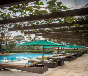 A poolside area with lounge chairs and green umbrellas, surrounded by greenery and a clear sky.