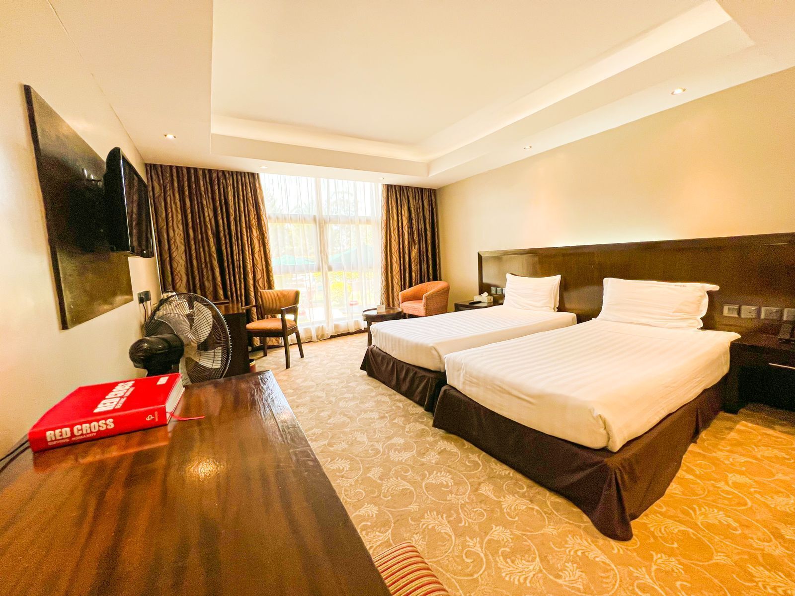 A hotel room with two beds, a desk, a chair, a fan, and a large window with curtains.