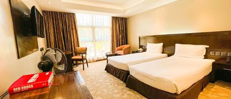 A hotel room with two beds, a desk, a chair, a fan, and a large window with curtains.