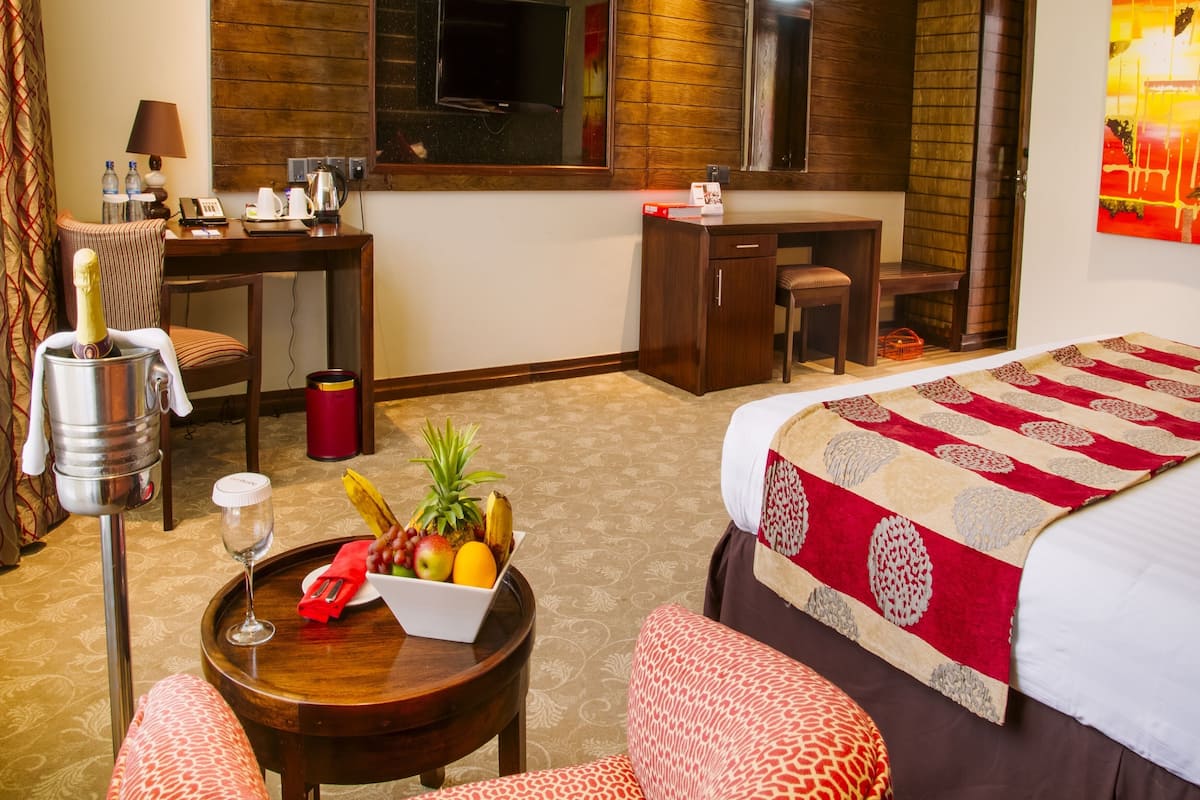 Habitación de hotel con cama, escritorio, silla, una mesita con frutero y copa de vino, y un espejo.