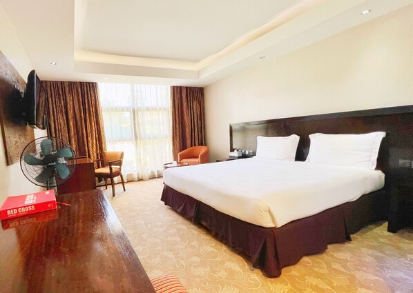 A hotel room with a large bed, a desk with a chair, a TV mounted on the wall, and a window with curtains.