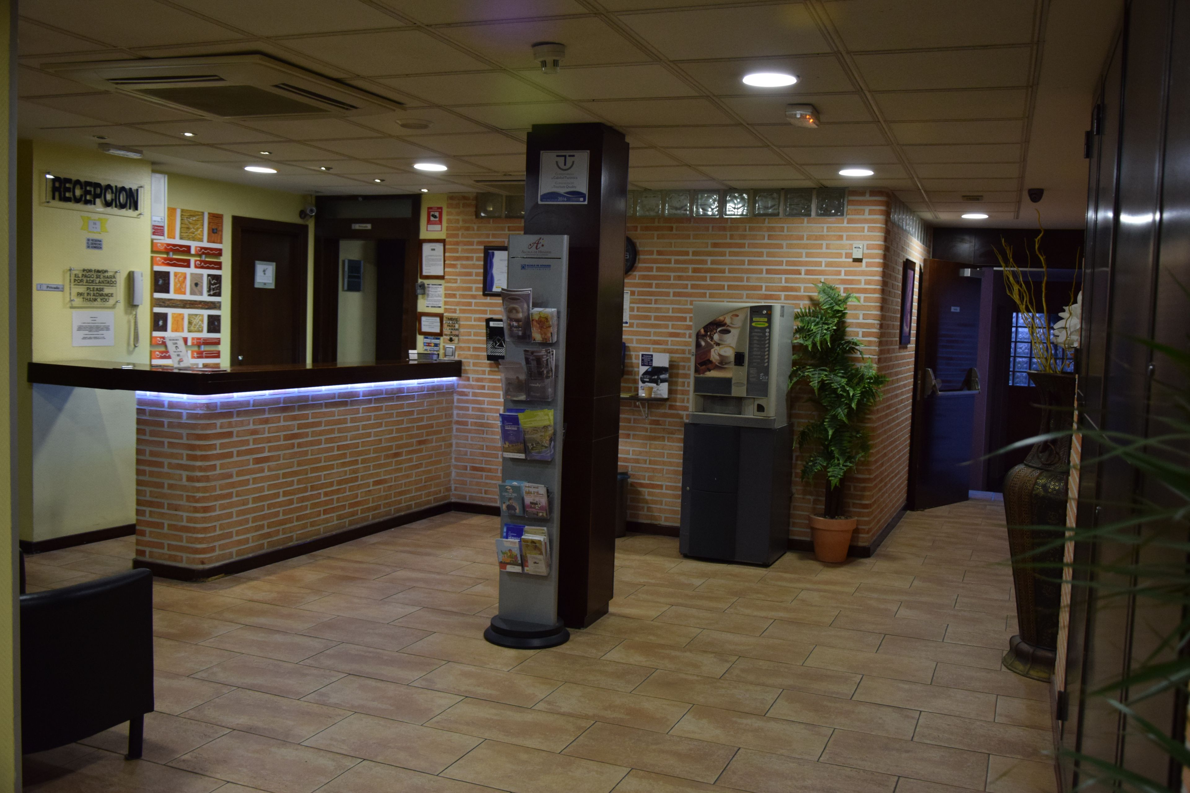 Zona de recepción con pared de ladrillo, mostrador de recepción, una columna con folletos informativos, máquina de café y planta en maceta.
