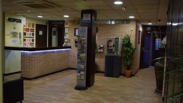 Zona de recepción con pared de ladrillo, mostrador de recepción, una columna con folletos informativos, máquina de café y planta en maceta.