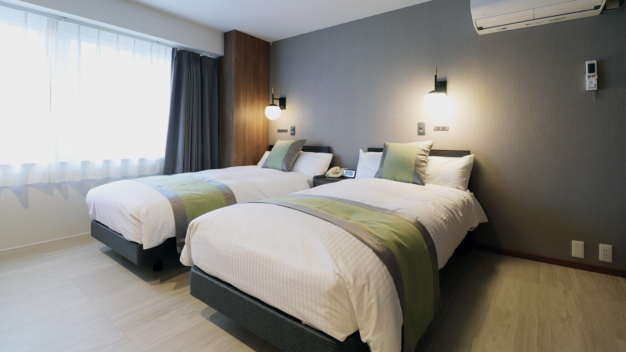 A hotel room with two beds, a window with curtains, a wall-mounted air conditioner, and a bedside table with a phone.