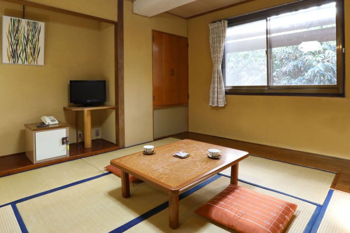Une pièce de style japonais traditionnel, avec un sol recouvert de tatamis, une basse table en bois entourée de coussins, un téléviseur posé sur un meuble et une armoire contenant un téléphone.