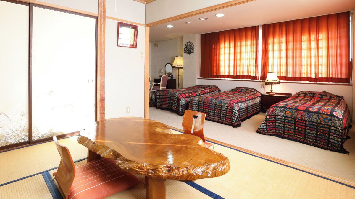 A room with tatami flooring, a wooden table, two beds with patterned bedding, a chair, and a desk with a lamp.
