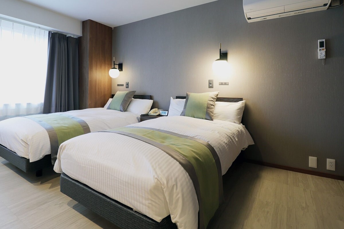 Une chambre d’hôtel avec deux lits, une fenêtre avec des rideaux, un climatiseur fixé au mur et une table de chevet avec un téléphone.