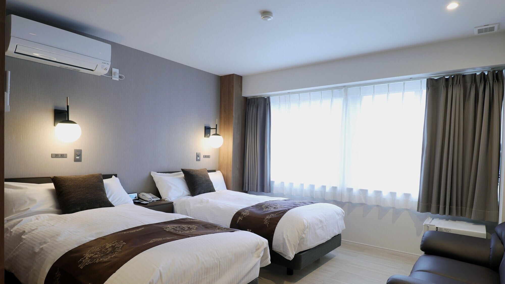 A hotel room with two beds, a desk, a chair, an air conditioning unit, and a window with curtains.