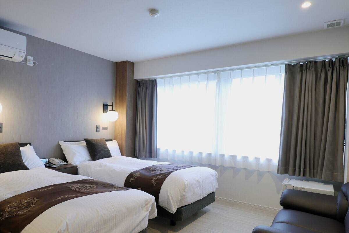 Une chambre d’hôtel avec deux lits, un bureau, une chaise, un appareil de climatisation et une fenêtre avec des rideaux.