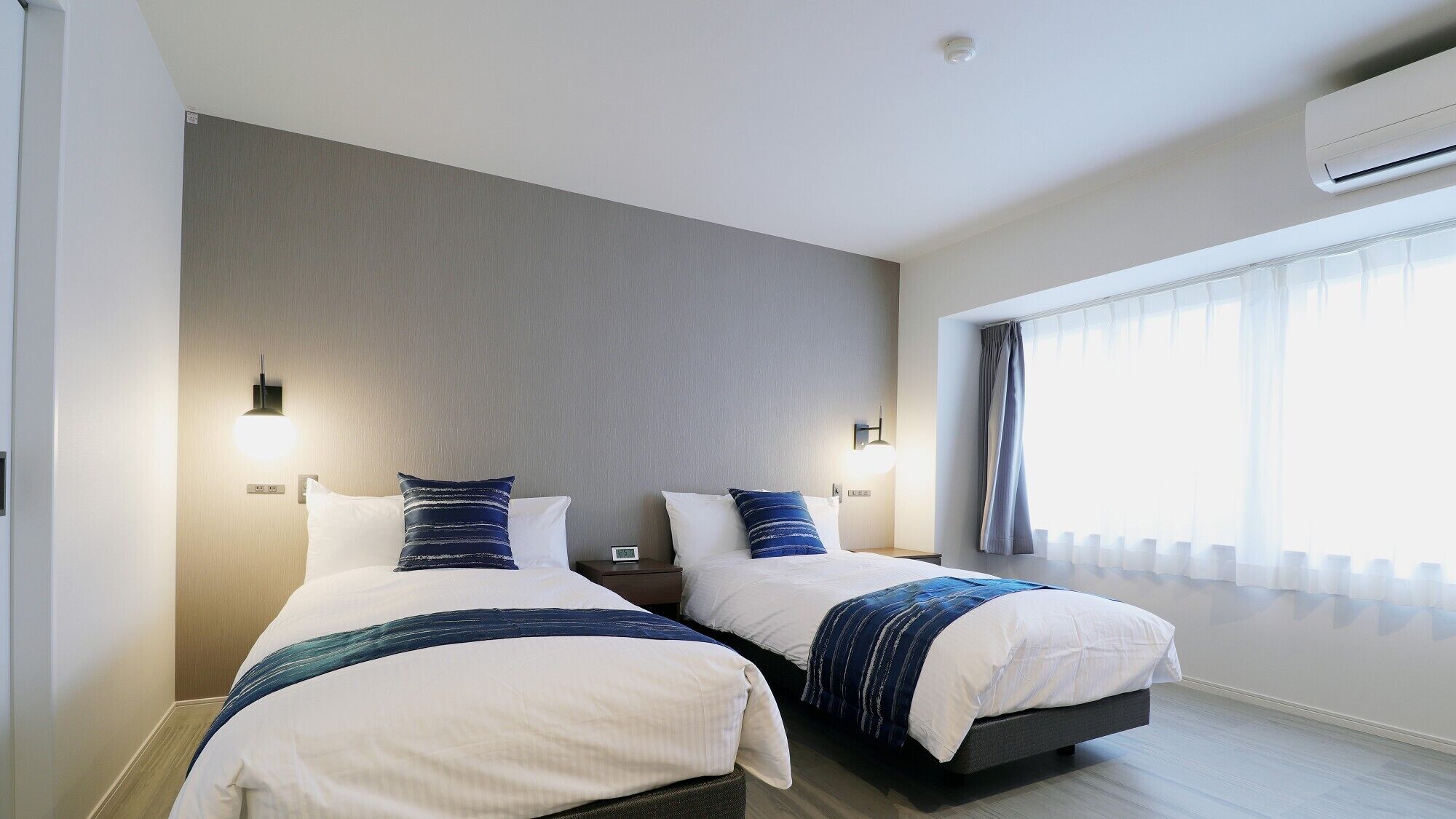 A hotel room with two beds, a large window with curtains, an air conditioning unit, and a wall-mounted light fixture.
