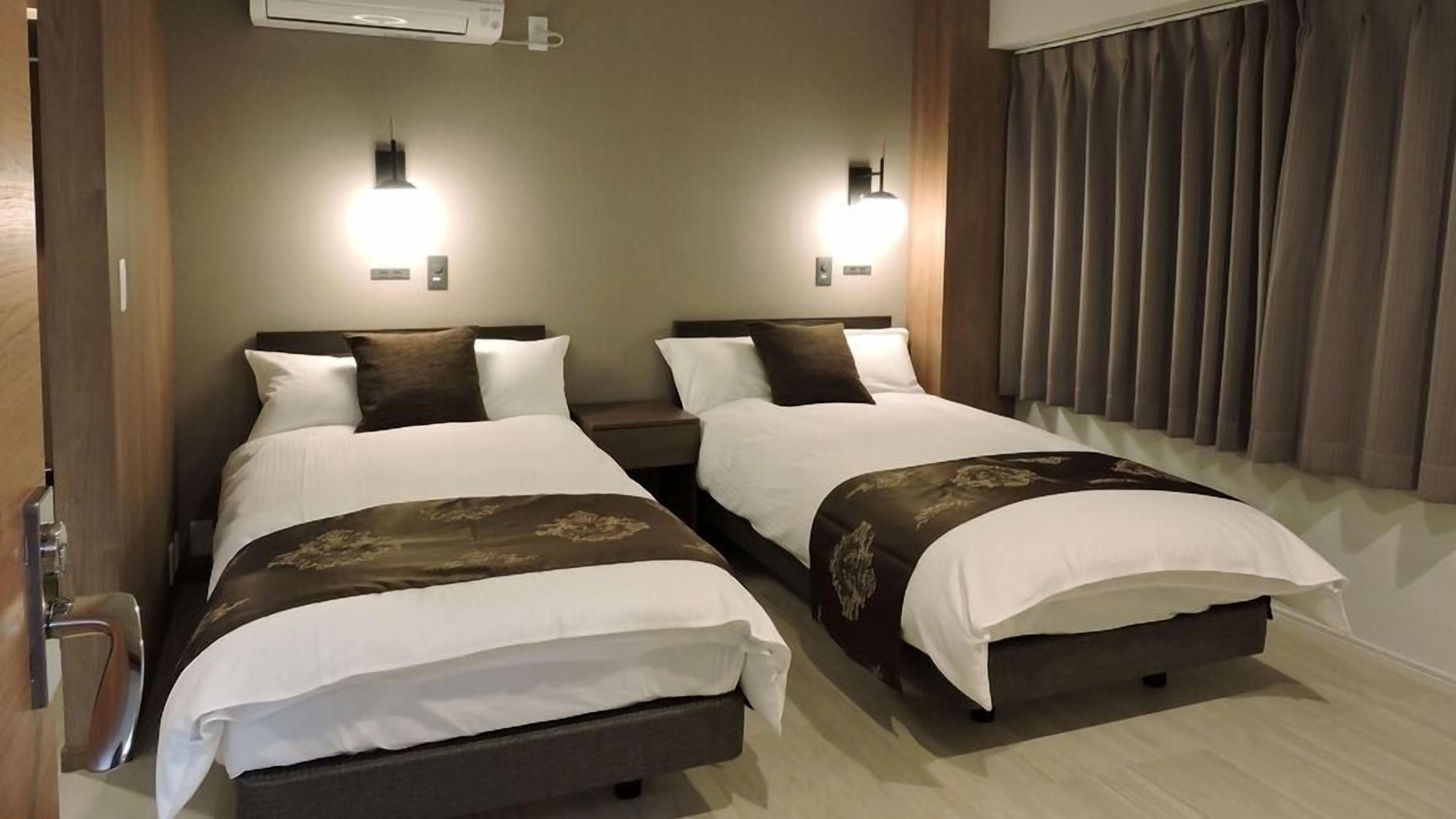 A hotel room with two beds, each with a headboard, bedside tables, and wall-mounted lights.