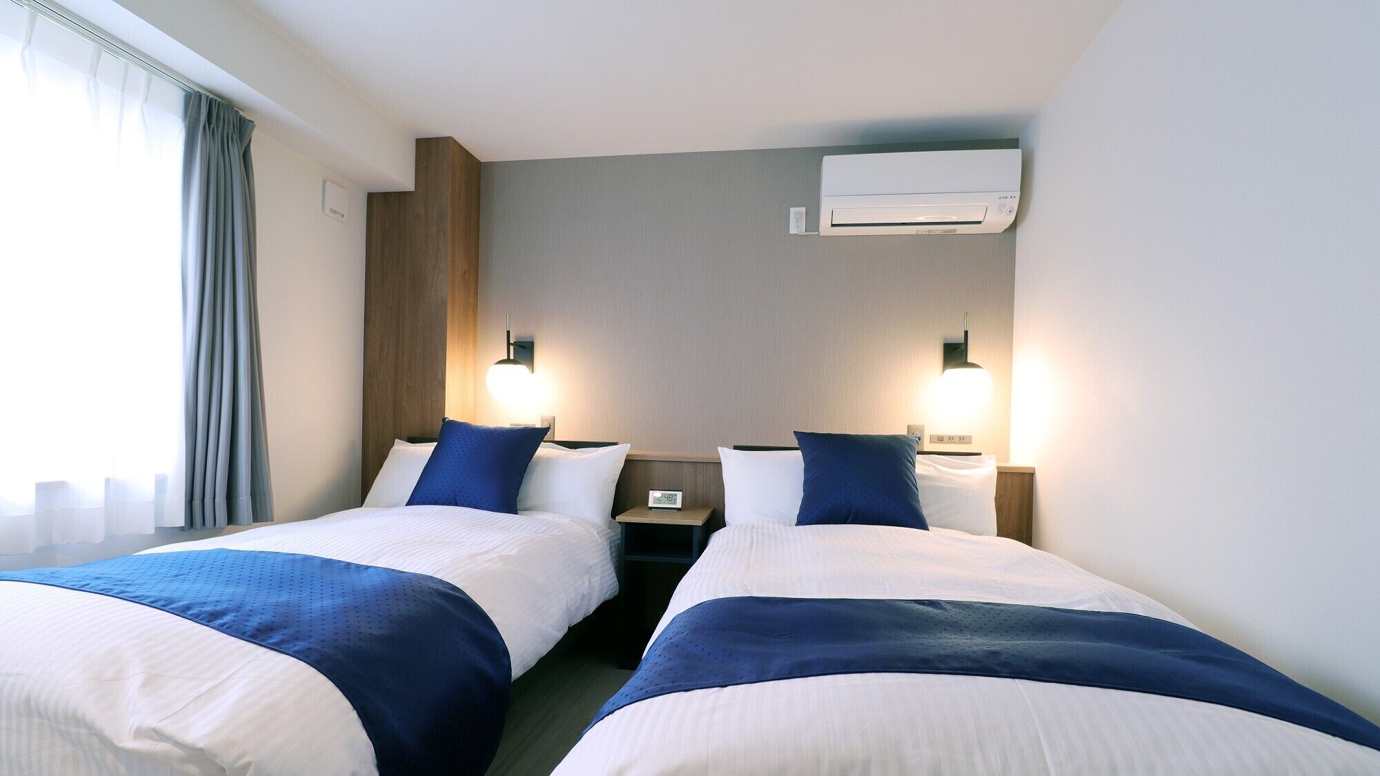 A hotel room with two beds, a nightstand with a clock, an air conditioning unit, and a window with curtains.