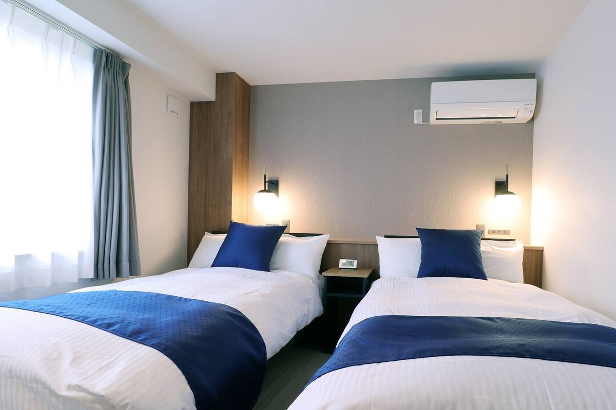 Une chambre d’hôtel avec deux lits, une table de chevet avec une horloge, un appareil de climatisation et une fenêtre avec des rideaux.
