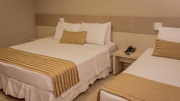 Quarto de hotel com duas camas, cada uma com roupa de cama branca e um cobertor bege riscado. Há uma mesinha de cabeceira com um telefone e uma luminária.