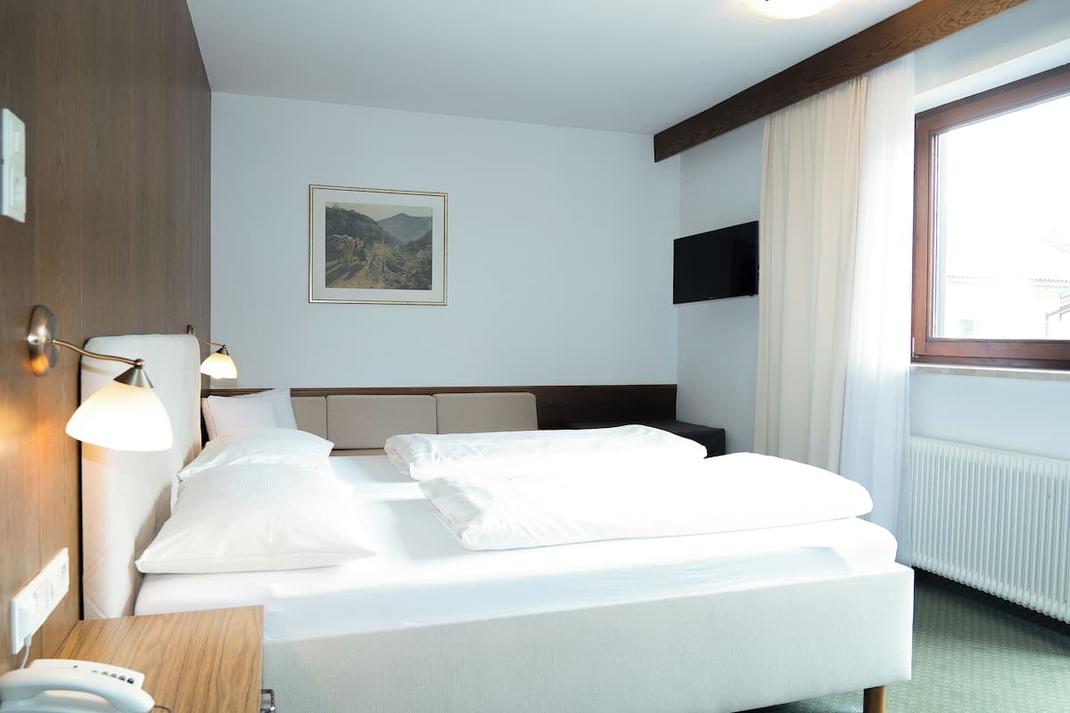 Habitación de hotel con cama, lámparas de noche, televisión montada, ventana con cortinas e imagen enmarcada en la pared.