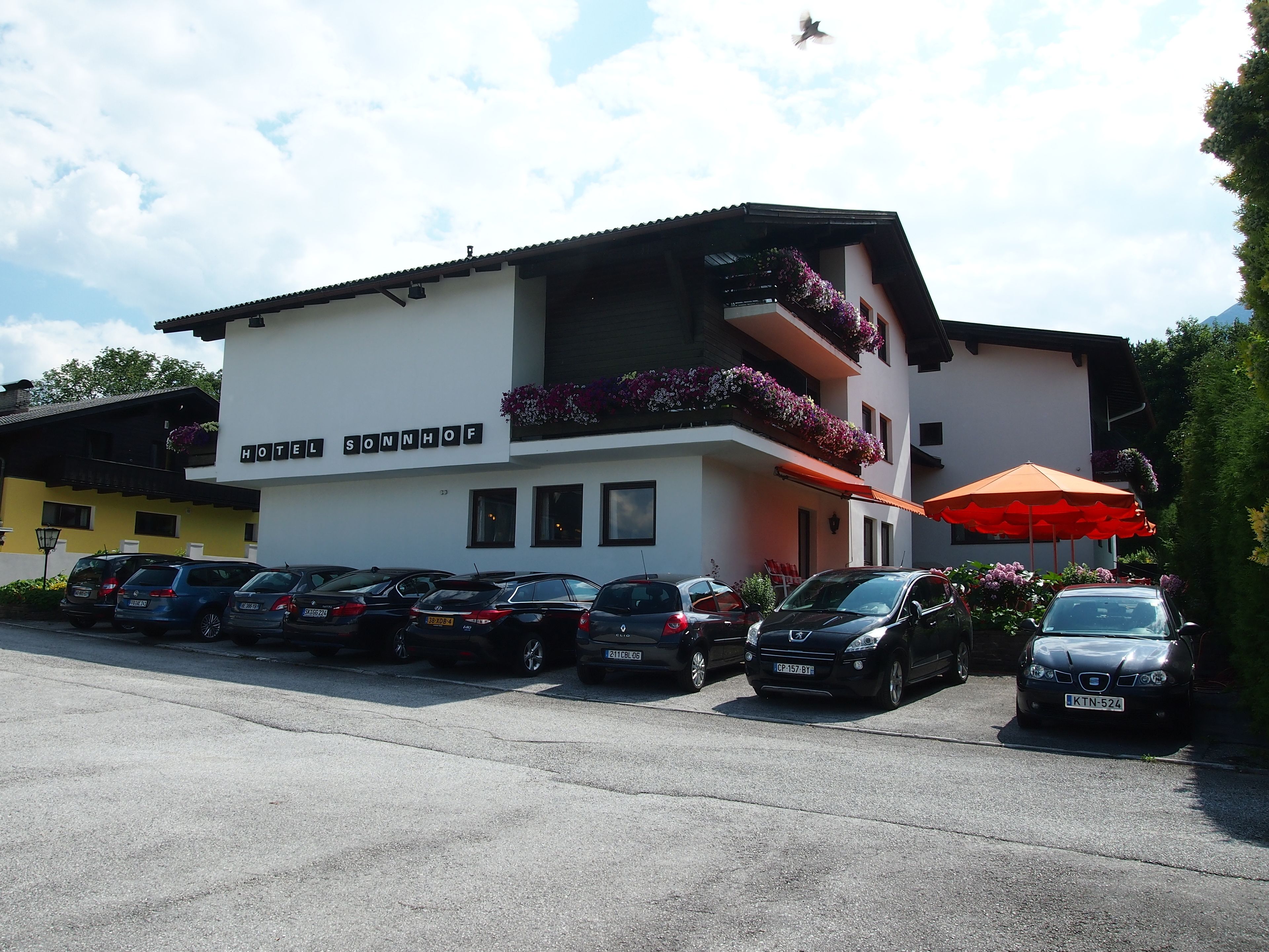 Un hotel con facciata bianca, balconi fioriti e un parcheggio con diverse auto.