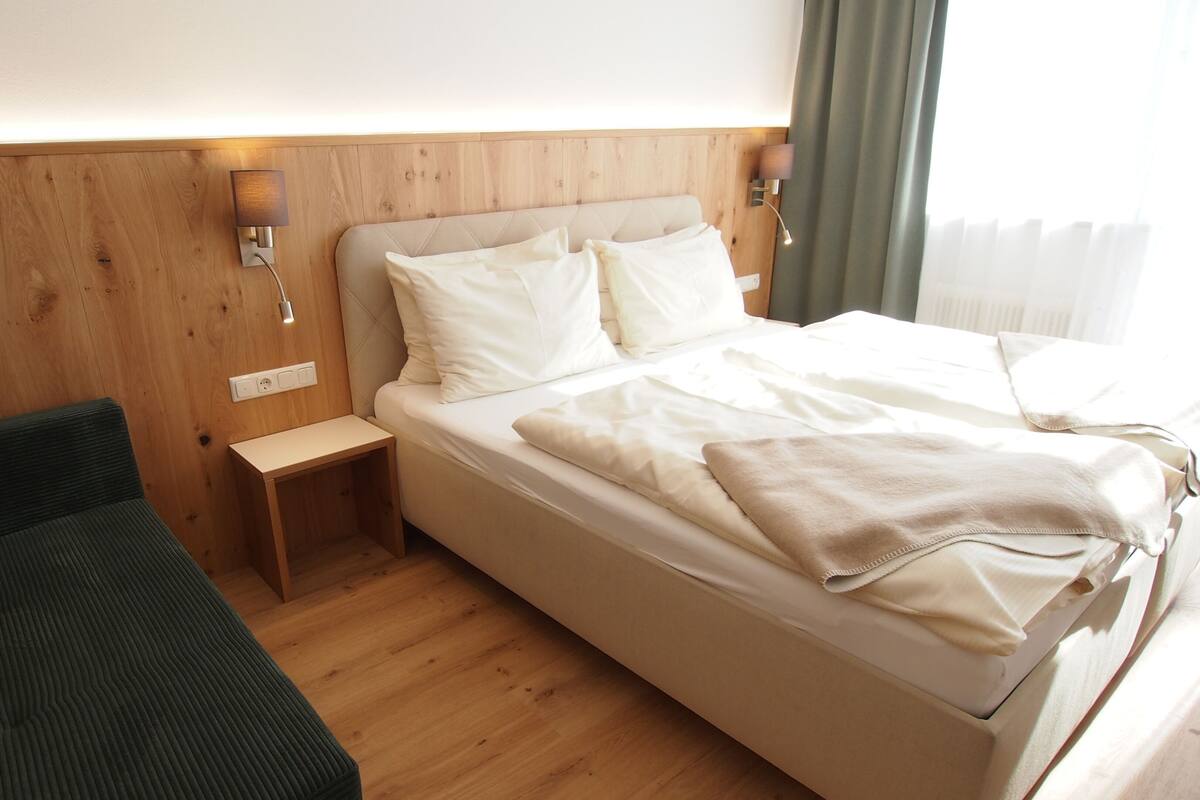 Una habitación de hotel con cama, mesita de noche, lámpara y una pequeña zona de estar.