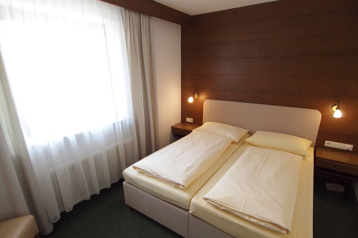 Cama doble con ropa de cama blanca, cabecera de madera, dos lámparas de noche y una ventana con cortinas traslúcidas.