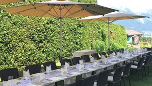 Une longue table à manger installée à l’extérieur, avec des chaises, des parasols et une haie en arrière-plan.