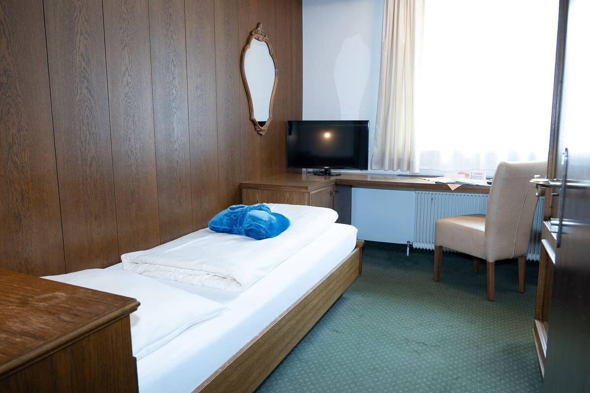 Habitación de hotel con pared de madera, cama con ropa blanca, escritorio con computadora y silla, y televisor.