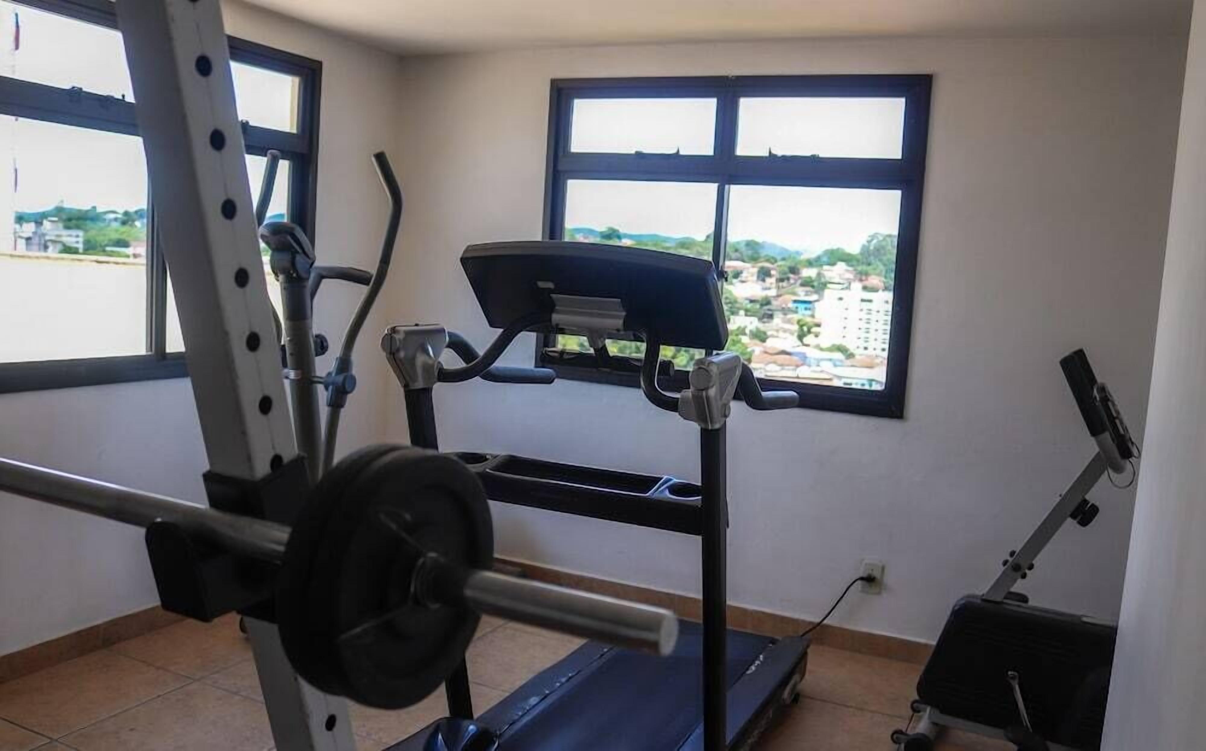 Et fitnessrum med træningsudstyr, herunder et løbebånd og en vægtbænk, med vinduer der giver udsigt til bygninger.