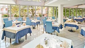 A restaurant with multiple tables, blue chairs, and a glass ceiling allowing natural light. - Hotel Europa (Grado)