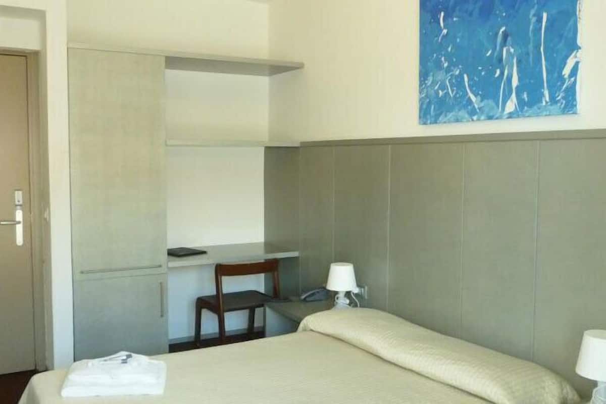 A hotel room with a bed, a desk, a chair, and a wall-mounted shelf.