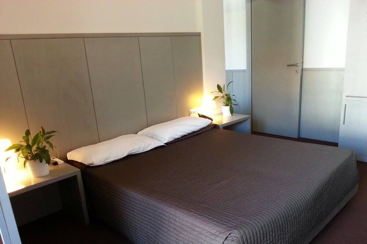 A neatly made bed with a headboard, two bedside tables with lamps, and a potted plant on one table.
