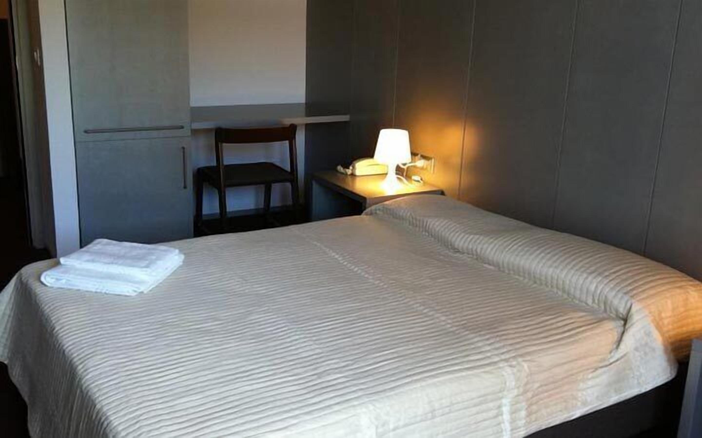A hotel room with a bed, a desk, a chair, and a bedside lamp.