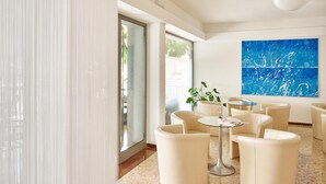A modern lounge area with a glass partition, a blue abstract painting, and a small round table with chairs. - Hotel Europa (Grado)
