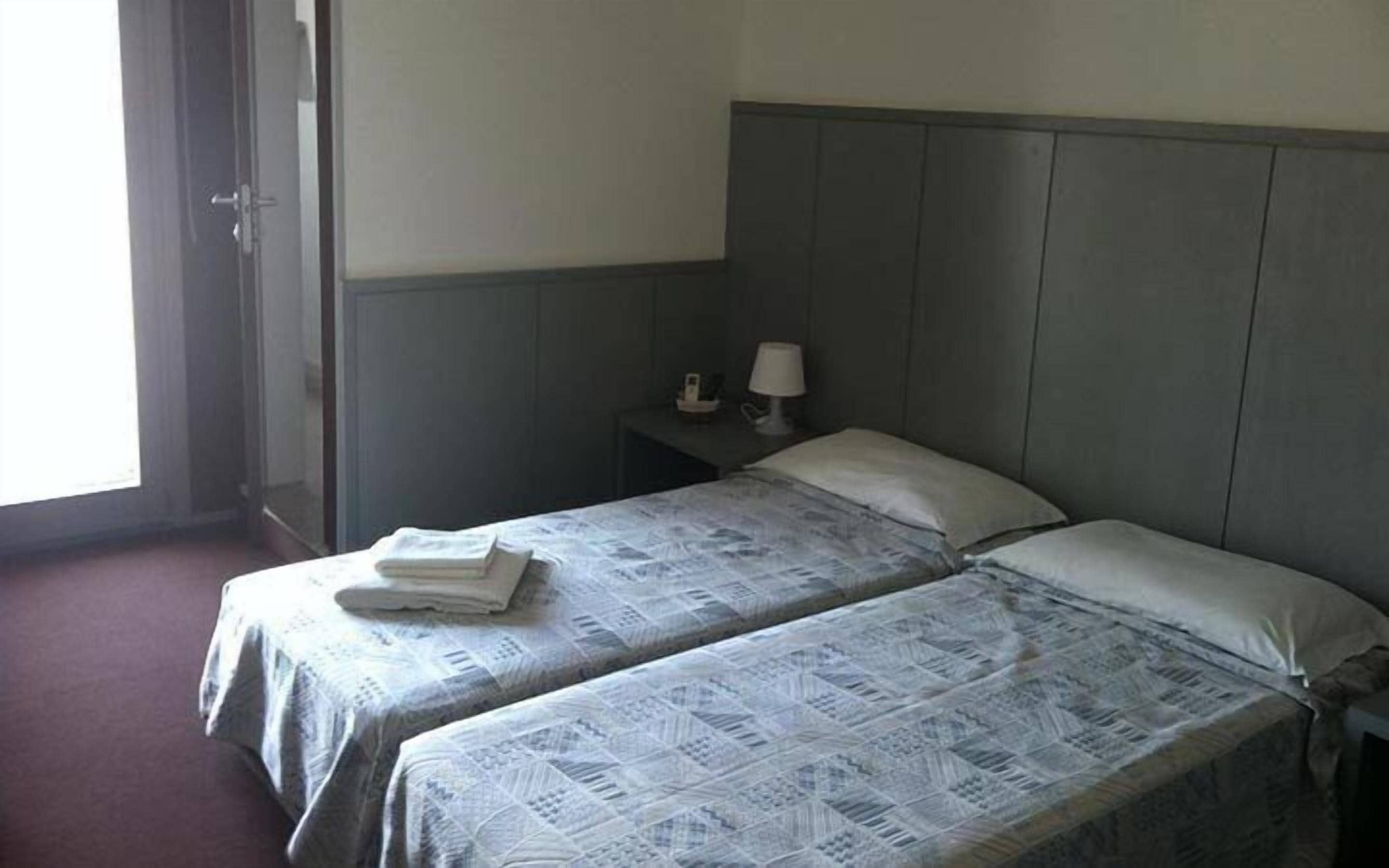 A small hotel room with two single beds, a nightstand with a lamp, and a window with curtains.