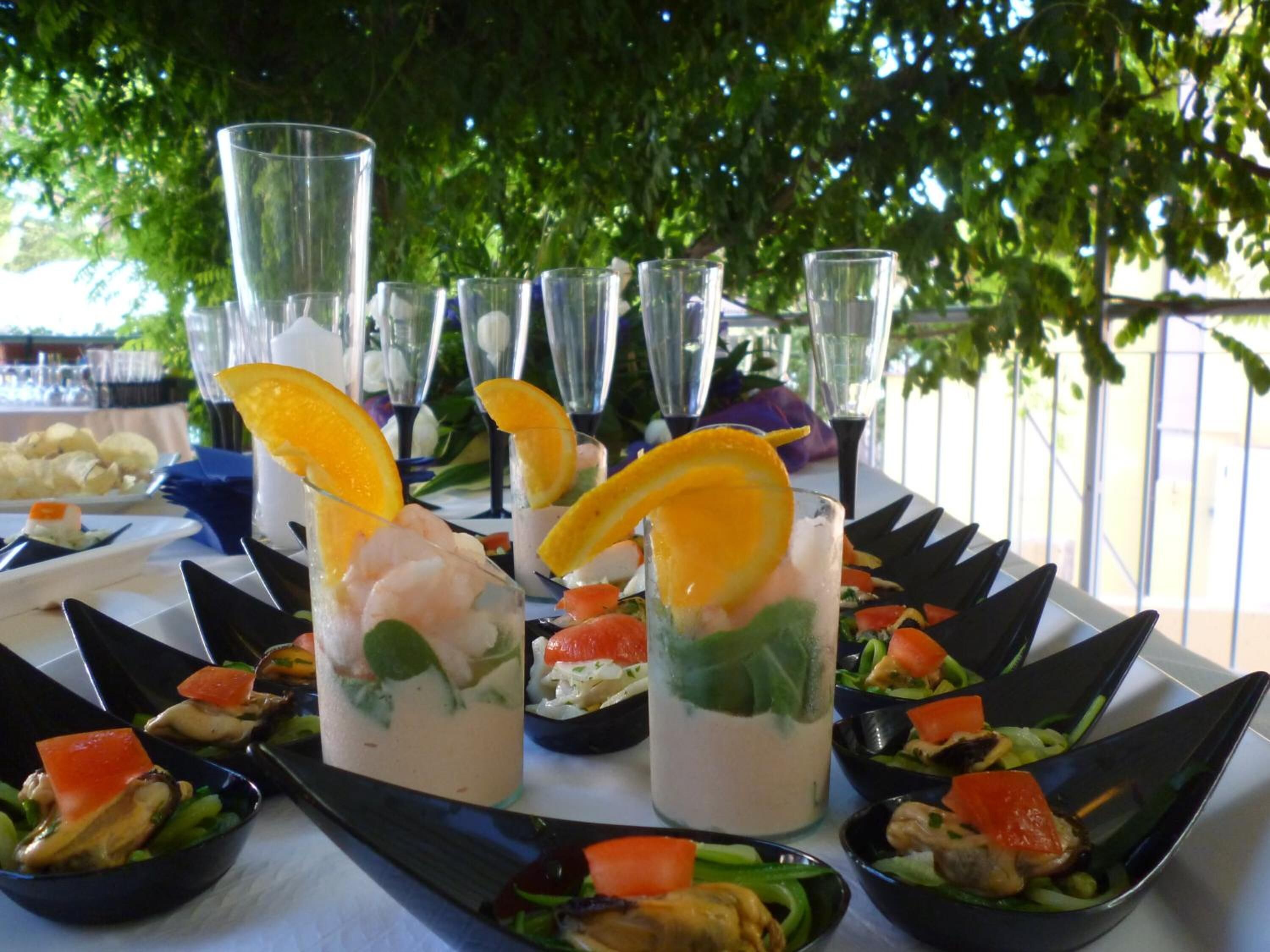 Mesa servida con diversos platos y bebidas, incluyendo mariscos y ensaladas, en un entorno al aire libre con árboles de fondo.