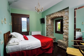 A bedroom with a bed, a stone wall, a wooden door, a window, a chair, and a chandelier.
