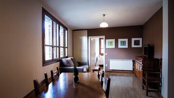 A dining area with a wooden table and chairs, a sofa, and a radiator. There are framed pictures on the wall and a television on a cabinet.