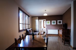 A dining area with a wooden table and chairs, a sofa, and a radiator. There are framed pictures on the wall and a television on a cabinet.